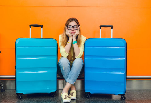Young girl with long hair in glasses is sitting  on orange background between two suitcases. She wears yellow sweater with jeans. She looks upset.