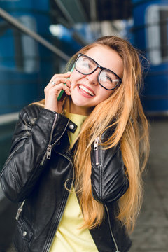 Close-up portrait of young girl with long hair in glasses outside in airport. She wears black jacket, speking on phone and smiling.