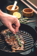 The cook lays a sprig of rosemary on an appetizing juicy beef steak, roasting in a pan for grilling.