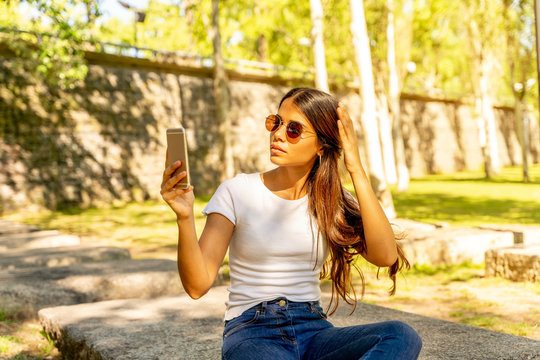 A beautiful young woman in a white shirt taking a selfie while sitting on a concrete block on a sunny day.