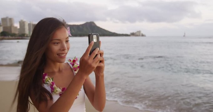 Happy young woman photographing using smartphone at Waikiki Beach. Smiling female tourist girl enjoying vacation at island in Honolulu wearing orchid lei garland taking photo.