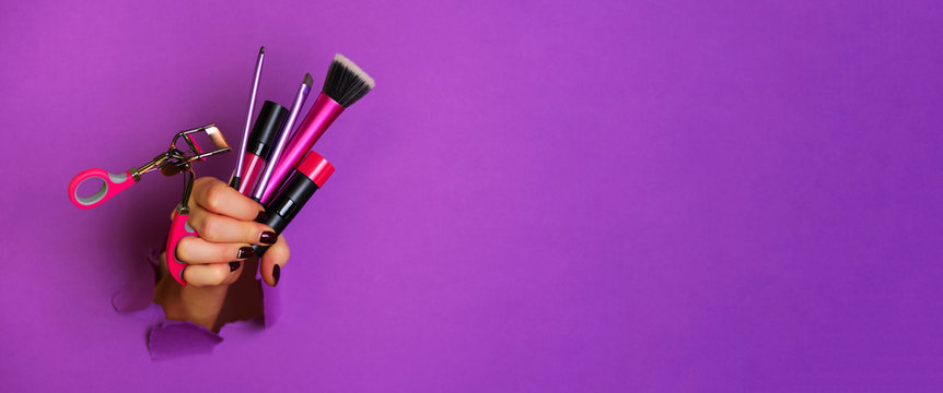 Woman hand with professional cosmetic tools for make up: brushes, mascara, lipstick, eyelash curler on violet background. Beauty concept. Banner for cosmetics sale. Copy space