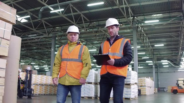 Workers walking and discussing stock inventory against tall shelves, in a warehouse with bright lamps.