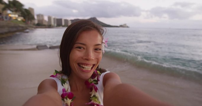 Asian woman taking selfie at Waikiki Beach. Smiling beautiful female tourist is wearing orchid lei garland during vacation. Girl puckering lips blowing kiss at camera in Honolulu.