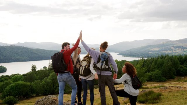 A multi ethnic group of five happy young adult friends cheer with arms in the air, high five and embrace at the summit during a mountain hike