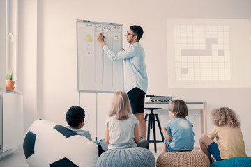 Young handsome teacher explaining project to group of kids sitting on poufs