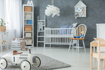 Chair next to bed under lamp in grey kid's bedroom interior with car toy on rug. Real photo