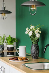 White roses in black and white vase next to coffee jug and big plate with cookies on wooden counter top