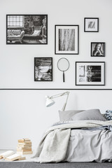 Books next to bed with white lamp in grey bedroom interior with gallery of photos. Real photo