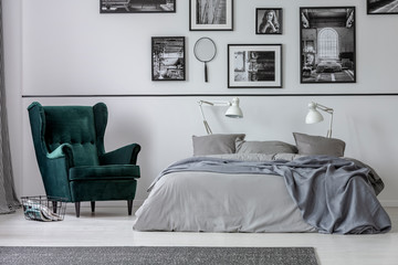 Armchair next to grey bed with white lamps in bedroom interior with gallery on the wall. Real photo