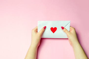 Female hand holding an envelope with a red heart. A love letter to the beloved. Valentines day concept. Greeting valentine card. Declaration of love. Wedding invitation. View from above. Flat lay