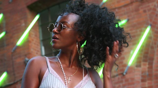 Fashionable young black woman wearing round sunglasses playing with her hair, smiling and lifting sunglasses, low angle, close up, blurred neon strip lights in background