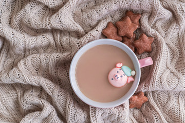 Cup of coffee with milk, marshmallow pig and star shaped ginger cookies on warm knitted beige blanket.