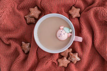 Cup of coffee with milk, marshmallow pig and star shaped ginger cookies on warm knitted coral blanket.