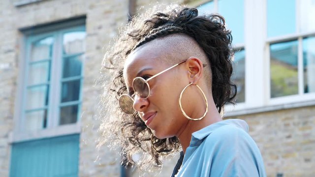 Fashionable young black woman wearing blue dress and sunglasses laughing to camera in the street, backlit, low angle, close up