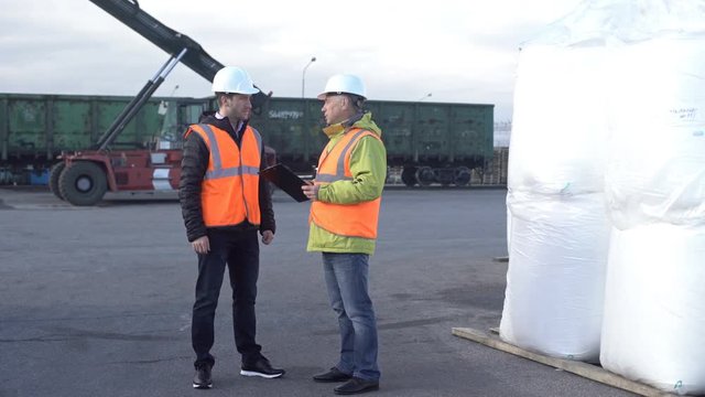 Two engineers wearing hardhats talking together while standing on a large commercial trainyard tracking inventory