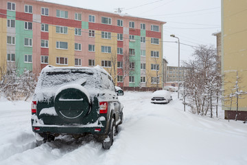 Off-road car on a snowy road in the yard after a snowfall. Around the snowdrifts and a lot of snow on the machines. Multi-colored panel buildings. Cold winter weather. Magadan, Far East of Russia.