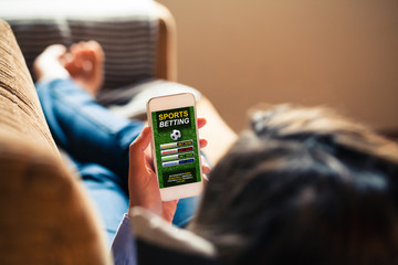 Woman holding a smart phone with sports betting website app in the screen, while lying down at home at home.