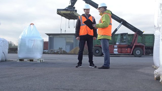 Two engineers wearing hardhats talking together while standing on a large commercial trainyard tracking inventory