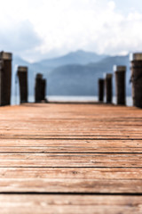 Naklejka na ściany i meble Wooden dock pier extending over blue lake water, mountains at lago di garda