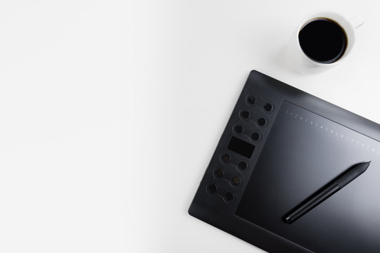 Top view on graphic designer's workspace. White table top with black graphic tablet, pen and cup of coffee. Minimalist flat lay. Illustrator's work place with copy space.