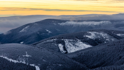 Beautiful view of wooden and snowy mountains partially covered in clouds with colorful sky, viewed...