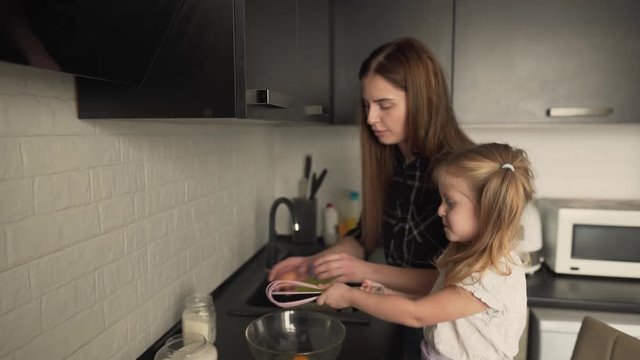 Mum and small daughter are standing in the kitchen, talking  and going to whisk eggs in a bowl. Family concept.