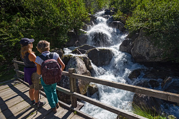 Two hikers are standing on a bridge to observe the rushing waters of a stream.