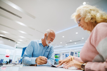 Grandparents looking at brochure and looking some products to buy while standing in tech store. Woman pointing at product she like.