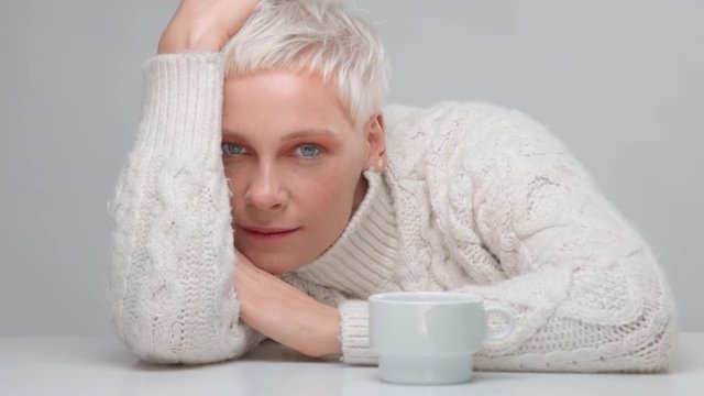 blonde woman with short haircut wears knitted sweater lyingo on the table with a white cup of hot dring behind