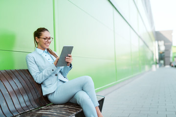 Beautiful Caucasian businesswoman sitting on the bench in front of business center and using tablet.
