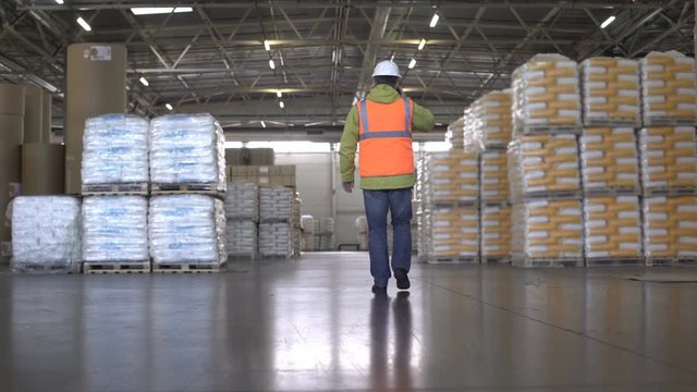 Worker in hard hat and orange uniform walking and talking by cellphone in warehouse - back view