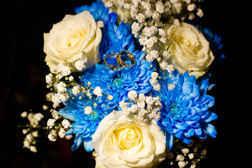 A pair of wedding gold rings on a bouquet of white and blue flowers on a black background. Shot close up.