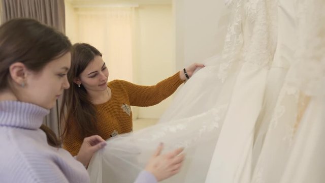 Two brunette girls are in wedding salon. One of them is future bride,another is her best friend. They are choosing dress for fitting.