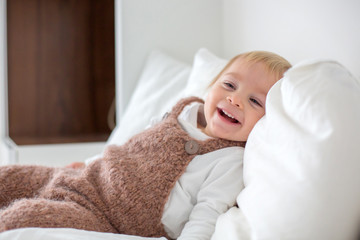 Sweet baby boy in cute overall, lying in bed, smiling with teddy bear stuffed toys, winter time