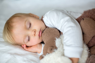 Sweet baby boy in cute overall, sleeping in bed with teddy bear stuffed toys, winter time