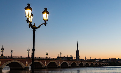 Fototapeta premium Street lights lit up at sunset, tram crossing Pont de Pierre bridge in the background, Bordeaux, France.