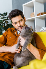 happy man with british shorthair cat sitting on sofa at home