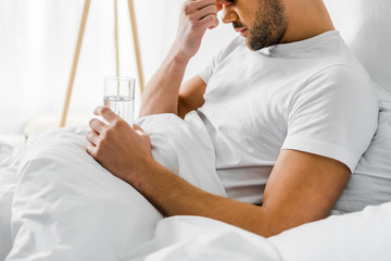 cropped view of man with headache holding glass of water in bed