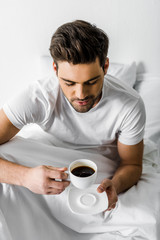 handsome smiling man in pajamas holding cup of coffee in bed