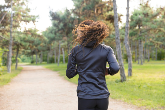 Image of fitness woman 20s wearing black tracksuit working out, while running through green park