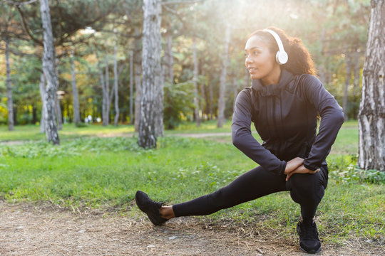 Photo of african american woman 20s wearing black tracksuit doing sports, and stretching body in green park