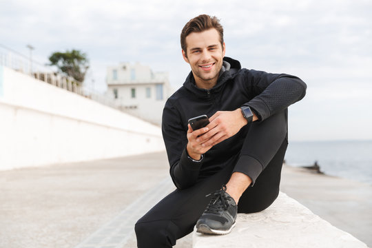 Image of muscular sportsman 30s in black sportswear, using smartphone while sitting on boardwalk at seaside