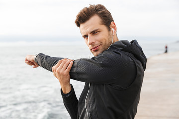 Fototapeta premium Image of european sportsman 30s in black sportswear and earphones, working out at seaside