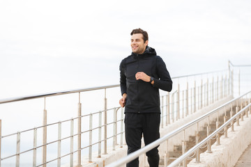 Image of satisfied sportsman 30s in black sportswear, running along pier at seaside