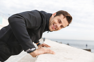 Image of cheerful man 30s in black sportswear, smiling while walking along boardwalk at seaside