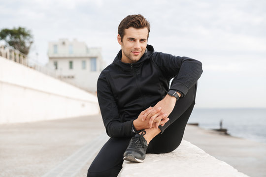 Image of sportive adult man 30s in black sportswear, sitting on boardwalk at seaside