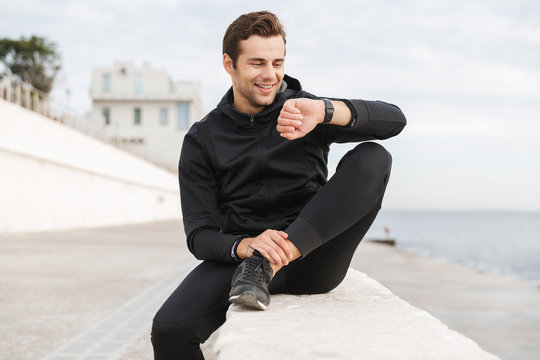 Image of handsome guy 30s in black sportswear looking at wrist watch, while sitting on boardwalk at seaside