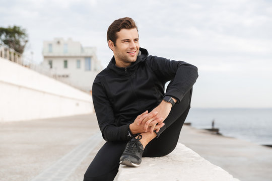 Image of muscular sportsman 30s in black sportswear, sitting on boardwalk at seaside