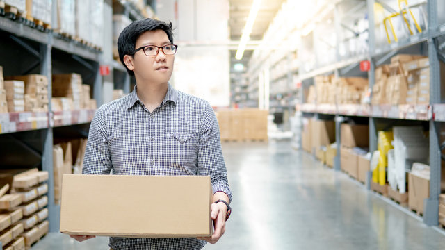 Young Asian man carrying cardboard box between row of shelves in warehouse. Shopping warehousing or working pick and packing concepts